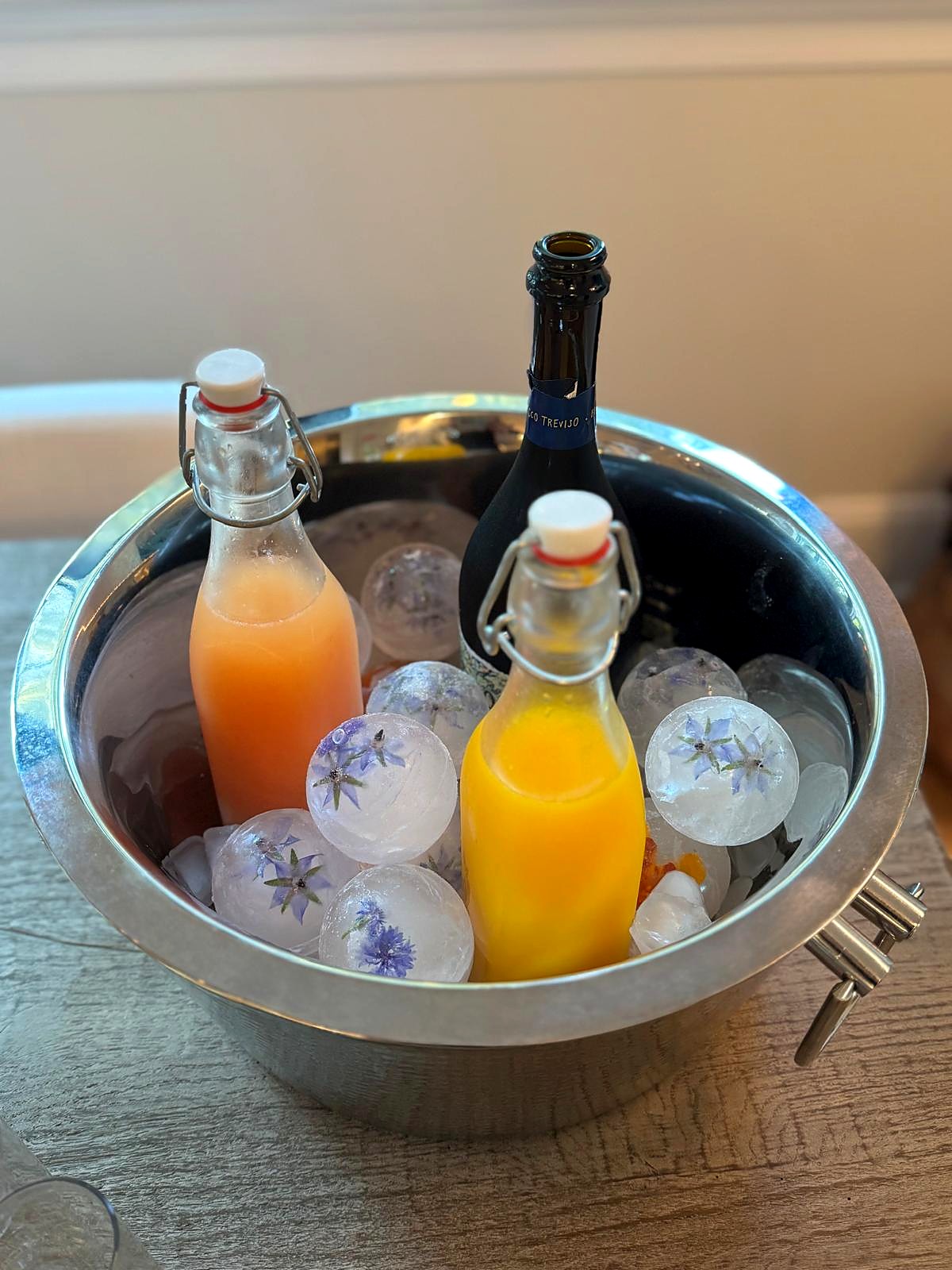 A chilled bucket of ice holding a bottle of Prosecco, two bottles of fresh-pressed juice, and ice spheres set with borage and cornflower