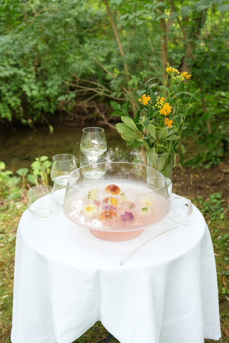 A garden table set with a punch bowl of botanical ice spheres beside a stream.