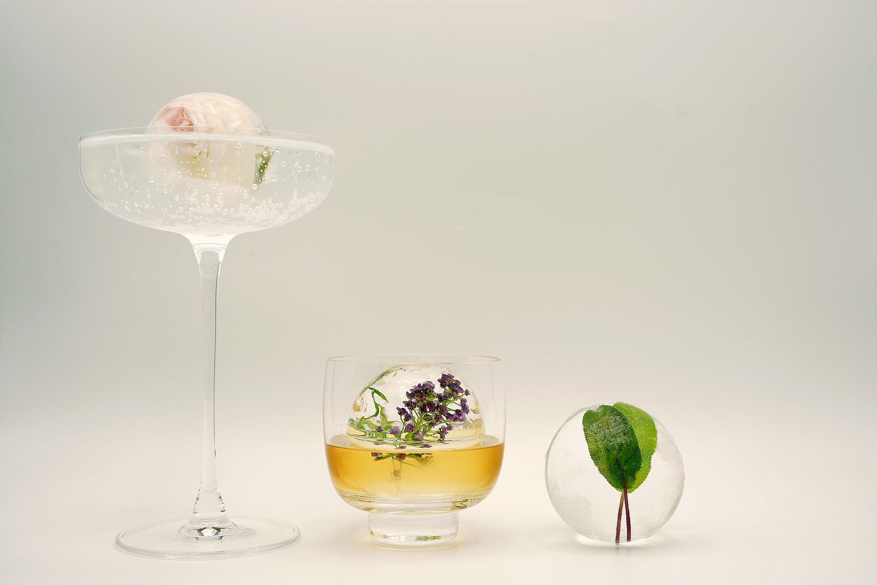 Three clear-ice spheres set with botanicals — a rose in a coupe, purple sweet alyssum in a glass of amber cocktail, and a sage leaf in ice.