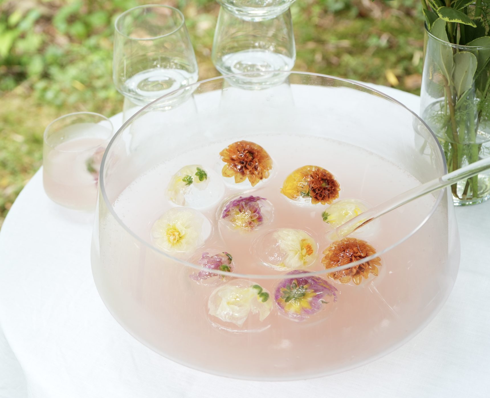 A glass punch bowl of clear-ice spheres set with edible blooms, served on a linen-draped table in a green garden.