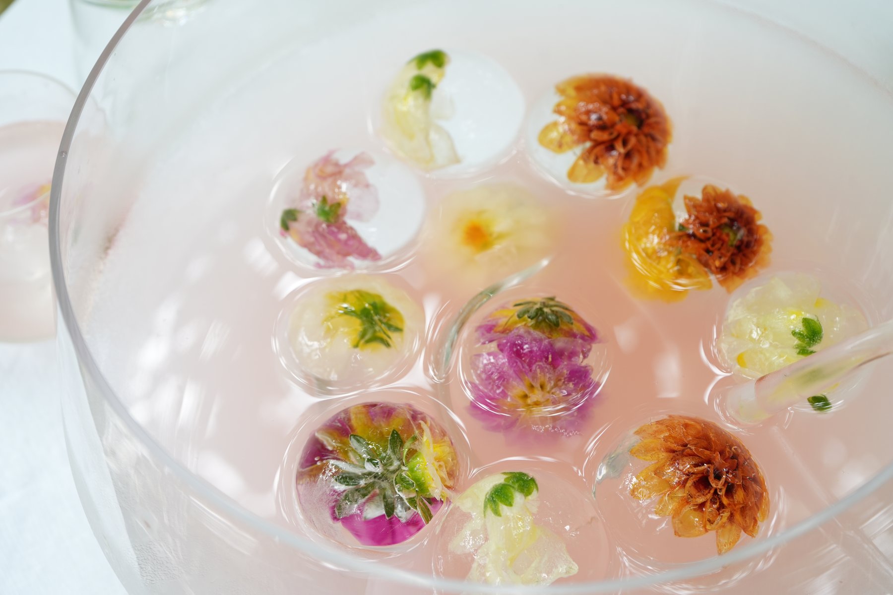 An overhead view of a bowl of clear-ice spheres, each set with a different edible bloom — dahlias, roses, violas, marigold, and greenery — floating in a pale pink punch.